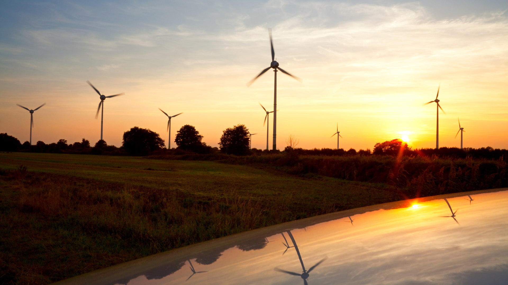 Champ d'éoliennes en fonctionnement au crépuscule, avec le soleil couchant en arrière-plan et les pales des turbines capturant l'énergie du vent. Le ciel est teinté d’orange et de rose, et les reflets des éoliennes apparaissent sur une surface brillante au premier plan, évoquant une transition énergétique respectueuse de l’environnement.