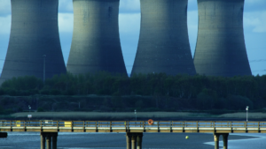 Quatre imposantes tours de refroidissement d’une centrale énergétique dominent l’arrière-plan, symboles d’une production électrique centralisée. Au premier plan, une passerelle surplombe une étendue d’eau paisible, contrastant avec l’infrastructure industrielle. Cette scène illustre les tensions et équilibres entre besoins énergétiques, territoires et mutations du mix énergétique.