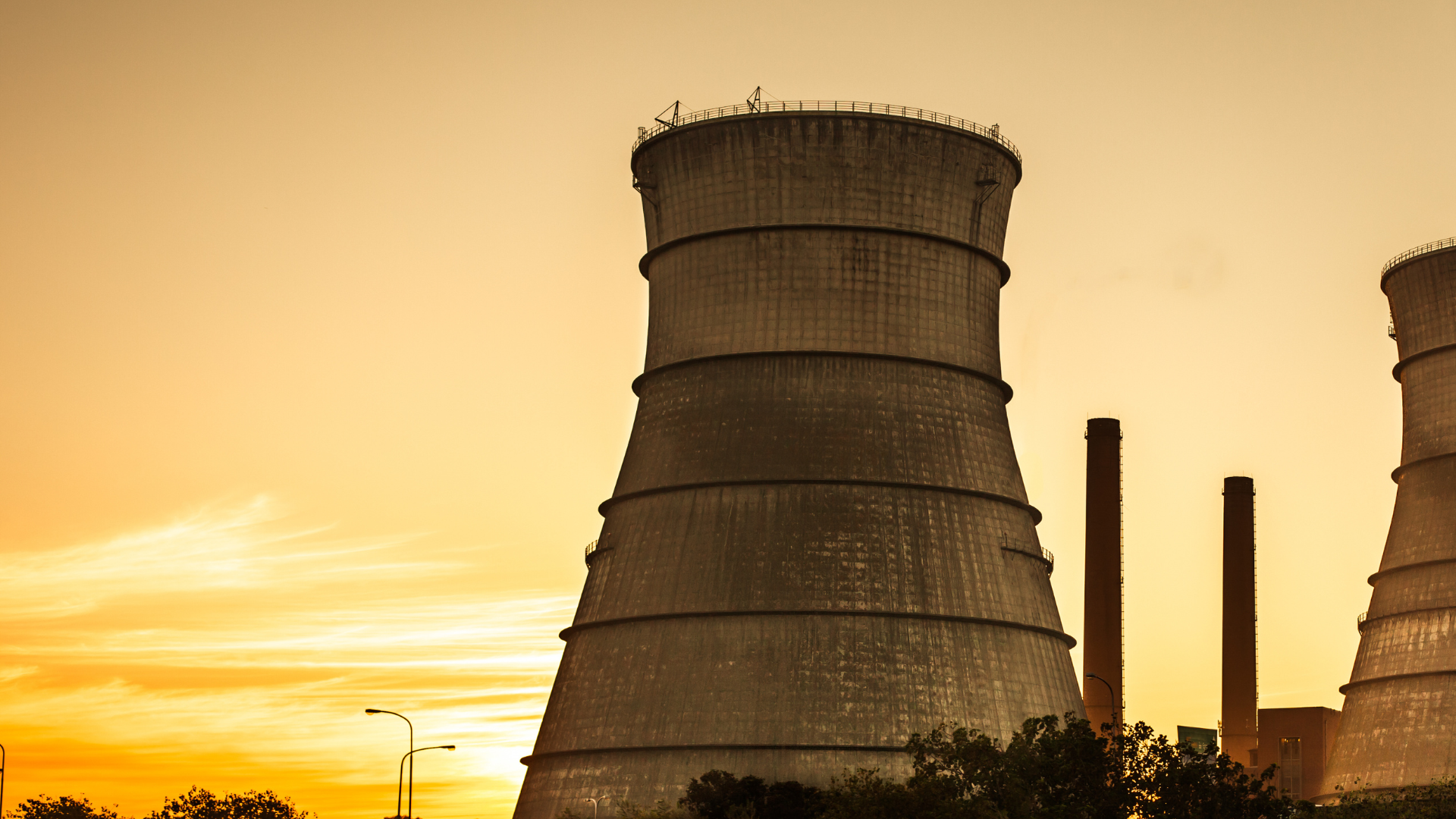 Tours de refroidissement d'une centrale électrique au coucher du soleil, avec le ciel orangé en arrière-plan et des cheminées visibles à droite.