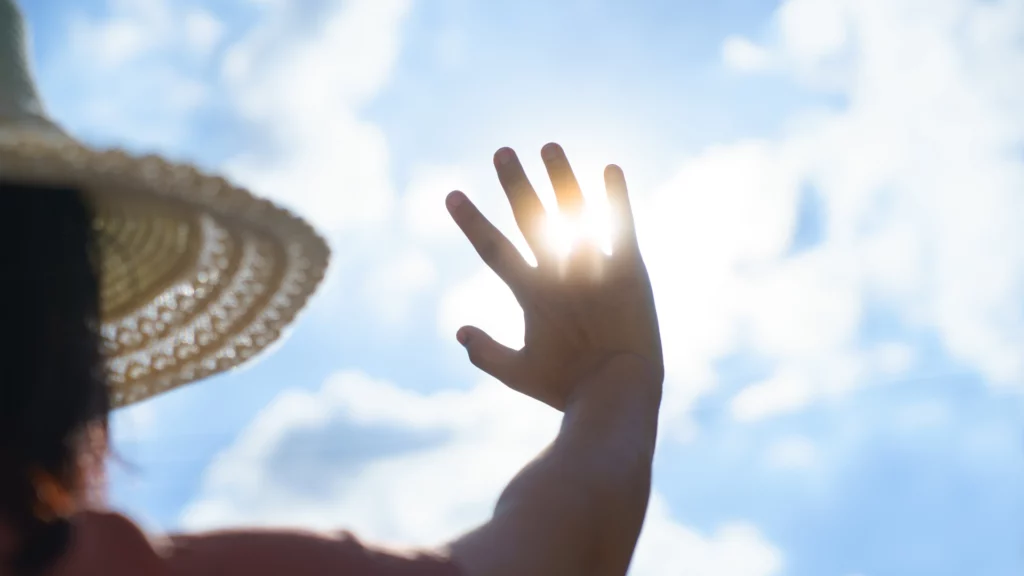 Personne portant un chapeau et levant la main pour se protéger du soleil, avec un ciel bleu en arrière-plan.