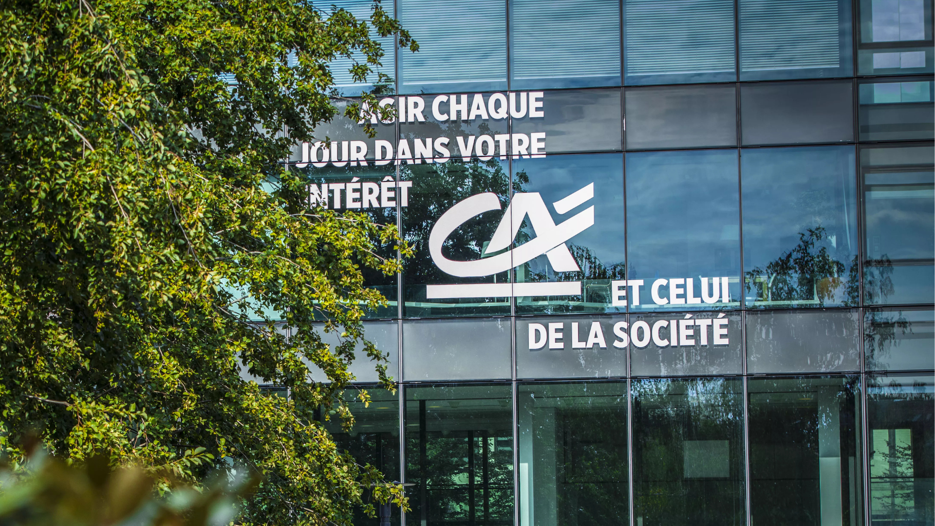 Façade vitrée d'un bâtiment Crédit Agricole avec logo et slogan, entourée d'arbres